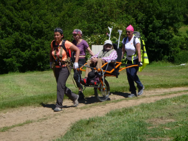 4 randonneurs pilotant une joëlette sur un petit chemin entouré d'herbe