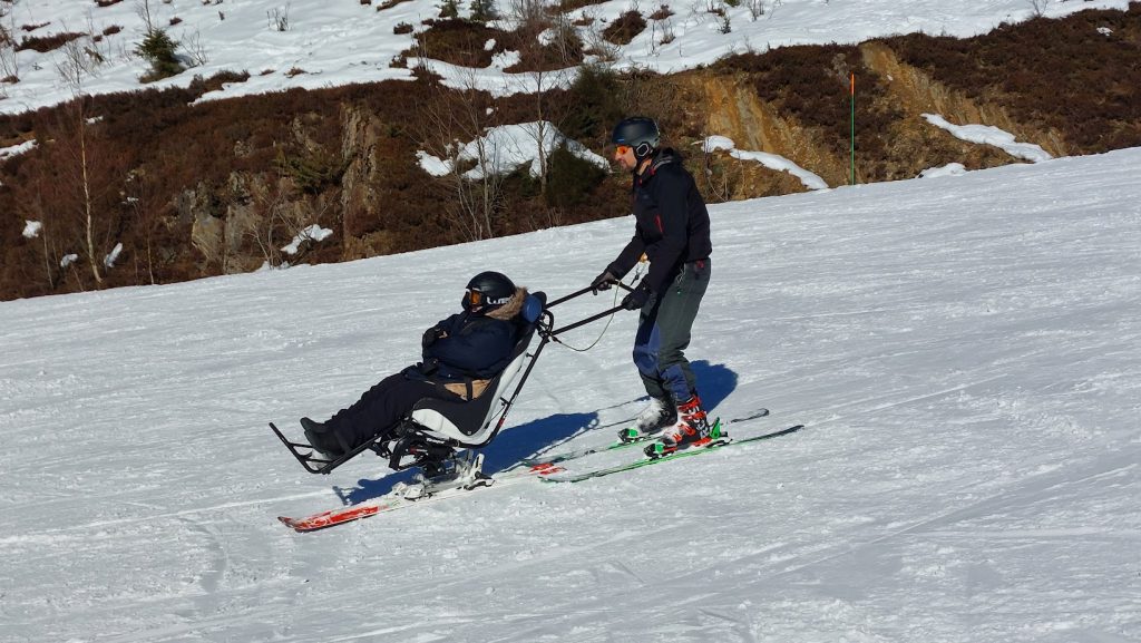 Descente de piste en fauteuil ski, un pilote debout et une personne en situation de handicap intellectuel sur le fauteuil.